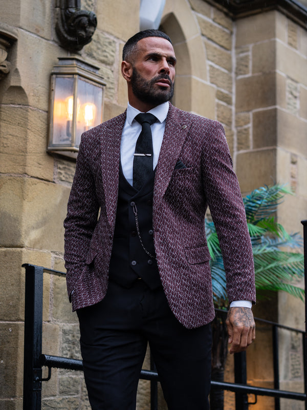 A man wearing a textured burgundy and black tweed blazer over a black vest, white shirt, and black tie, standing in front of a stone building.